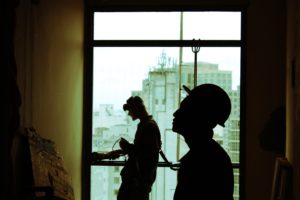 Two construction workers are in shadows, looking at a wall and standing in the light of a large window.