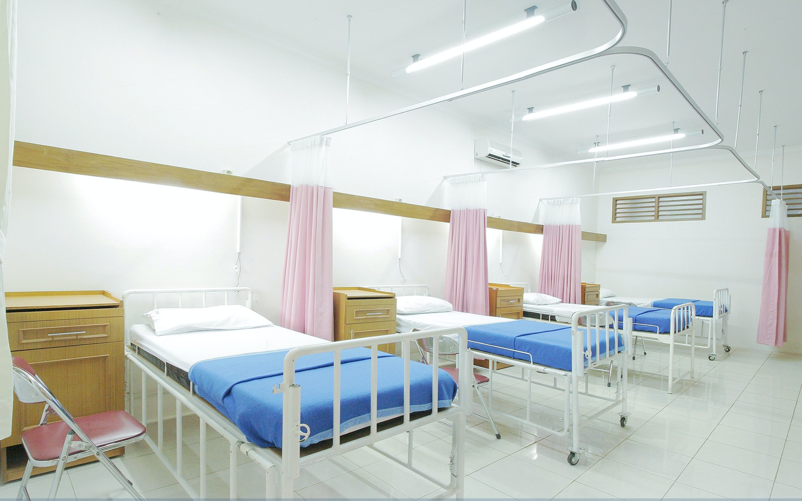Three hospital beds sit in a row surrounded by bright lights and white wallpaper.