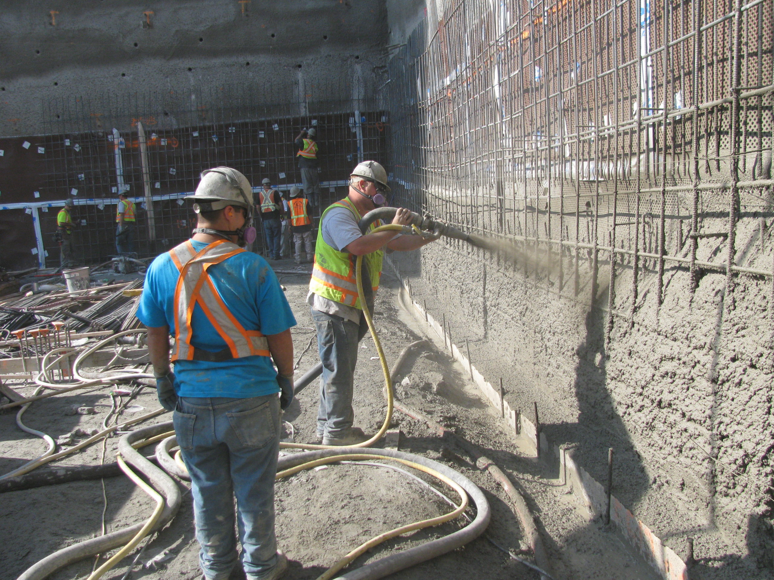 A construction worker looks at a coworker spraying Hard-Cem shotcrete against a barrier.