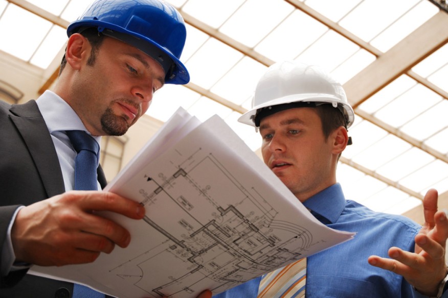 Two construction workers in hard hats and business suits look at some papers.