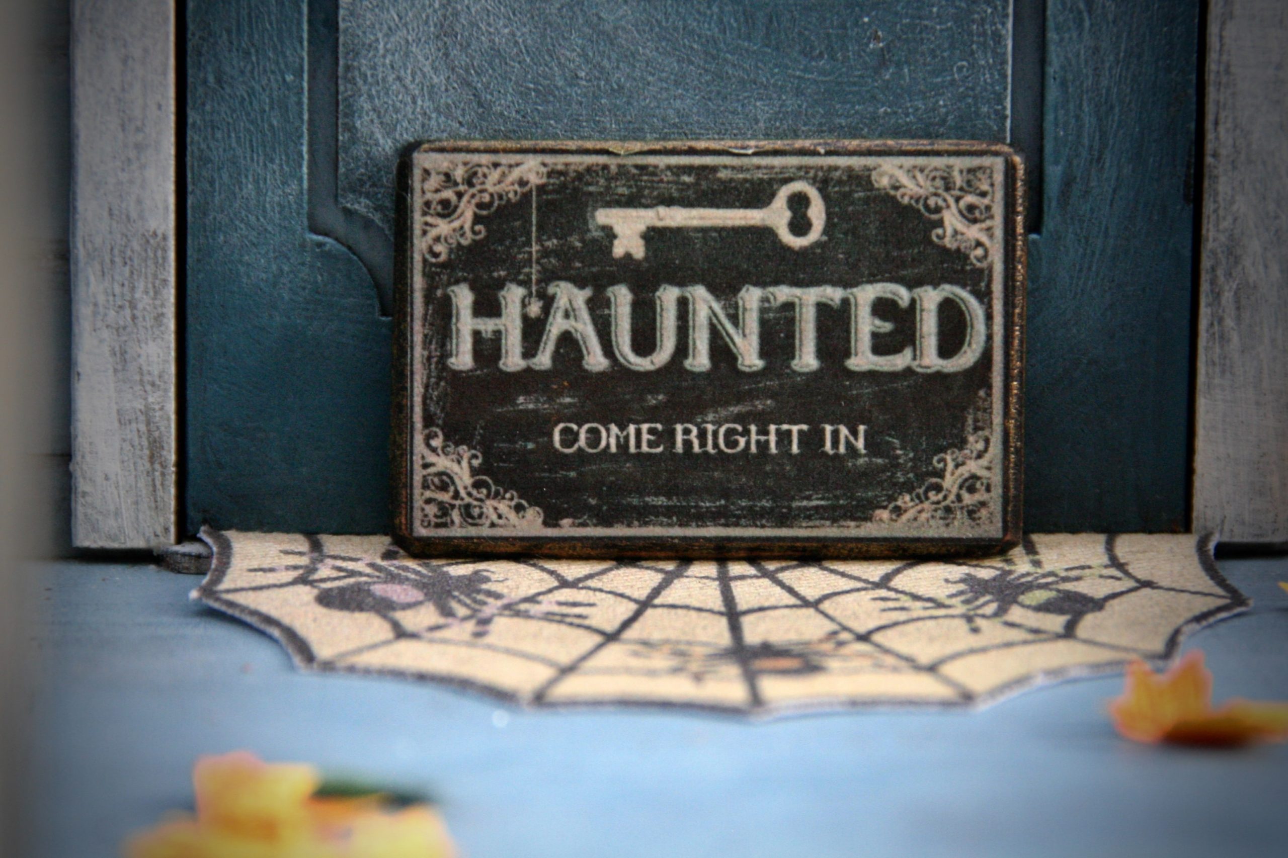 A sign resting against a door says "Haunted, Come Right In" and above the writing is an illustration of a key.