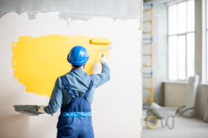A workperson is painting yellow over a white wall with ragged gray coloring at the top.