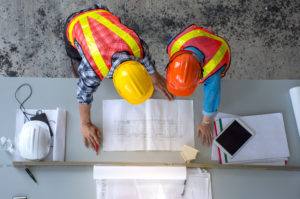 Two engineers with hard hats and safety vests on are looking at a blueprint that's spread out on a table.