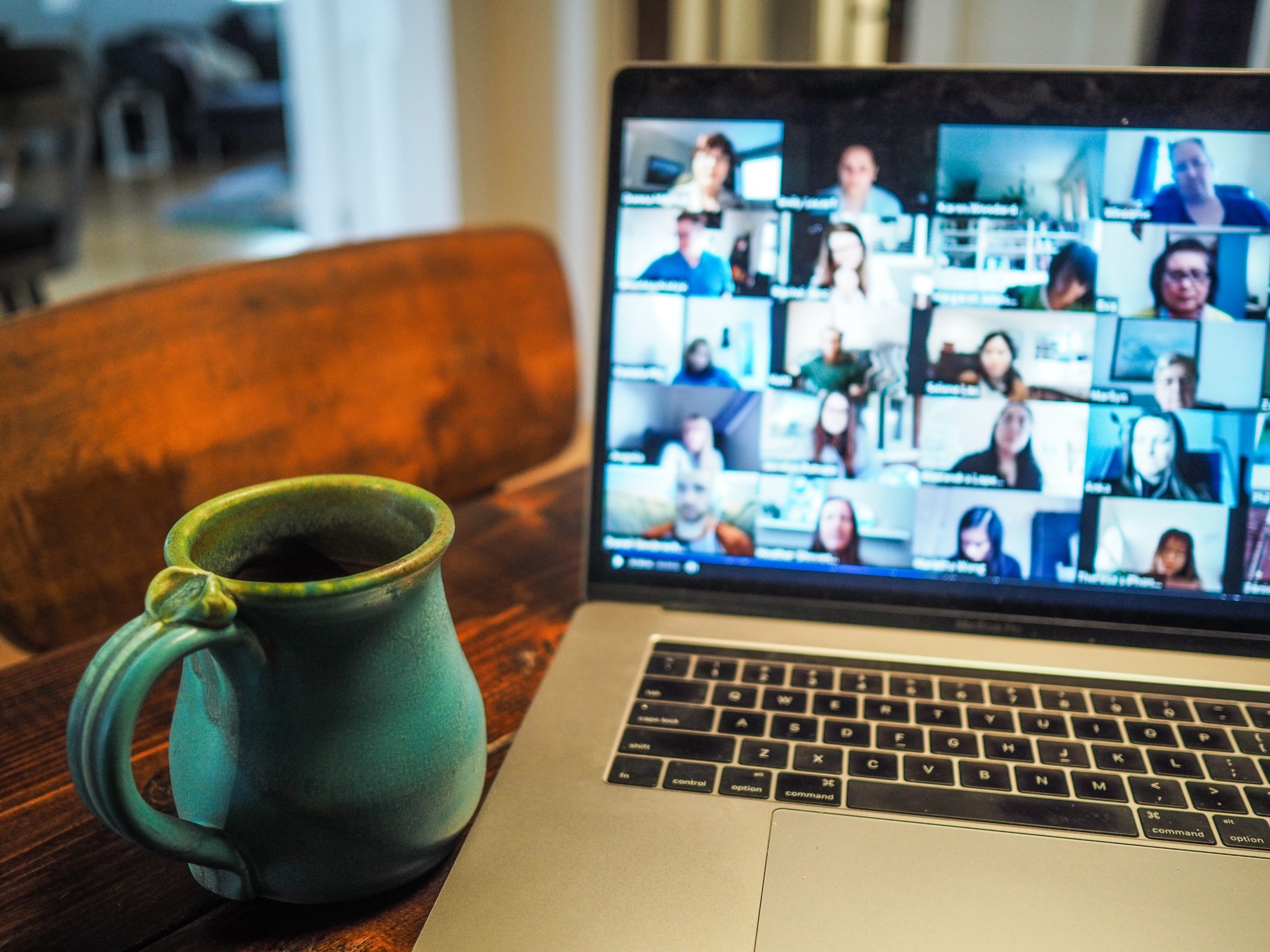 Several people can be seen on a laptop screen, participating in an online meeting, while a green mug of coffee sits to the left of the laptop.