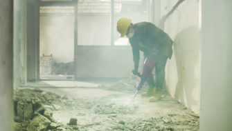 Dust surrounds a construction worker wearing a mask as they drill into a floor.