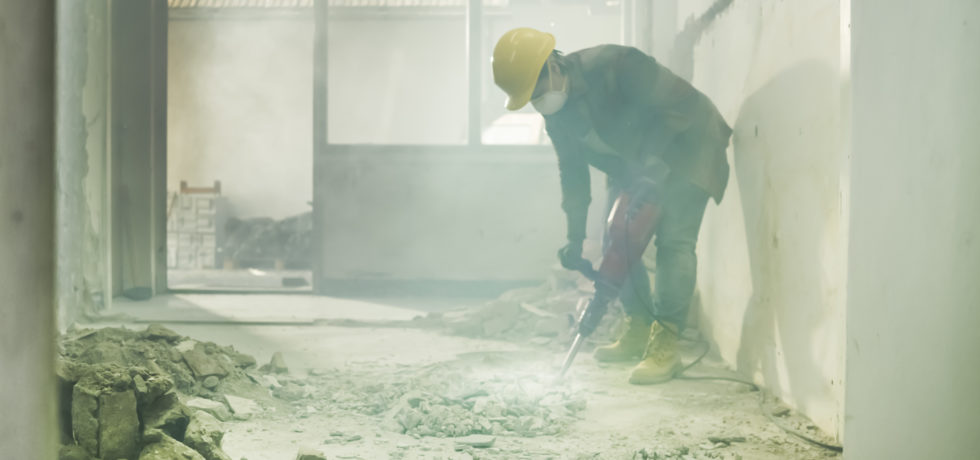 Dust surrounds a construction worker wearing a mask as they drill into a floor.