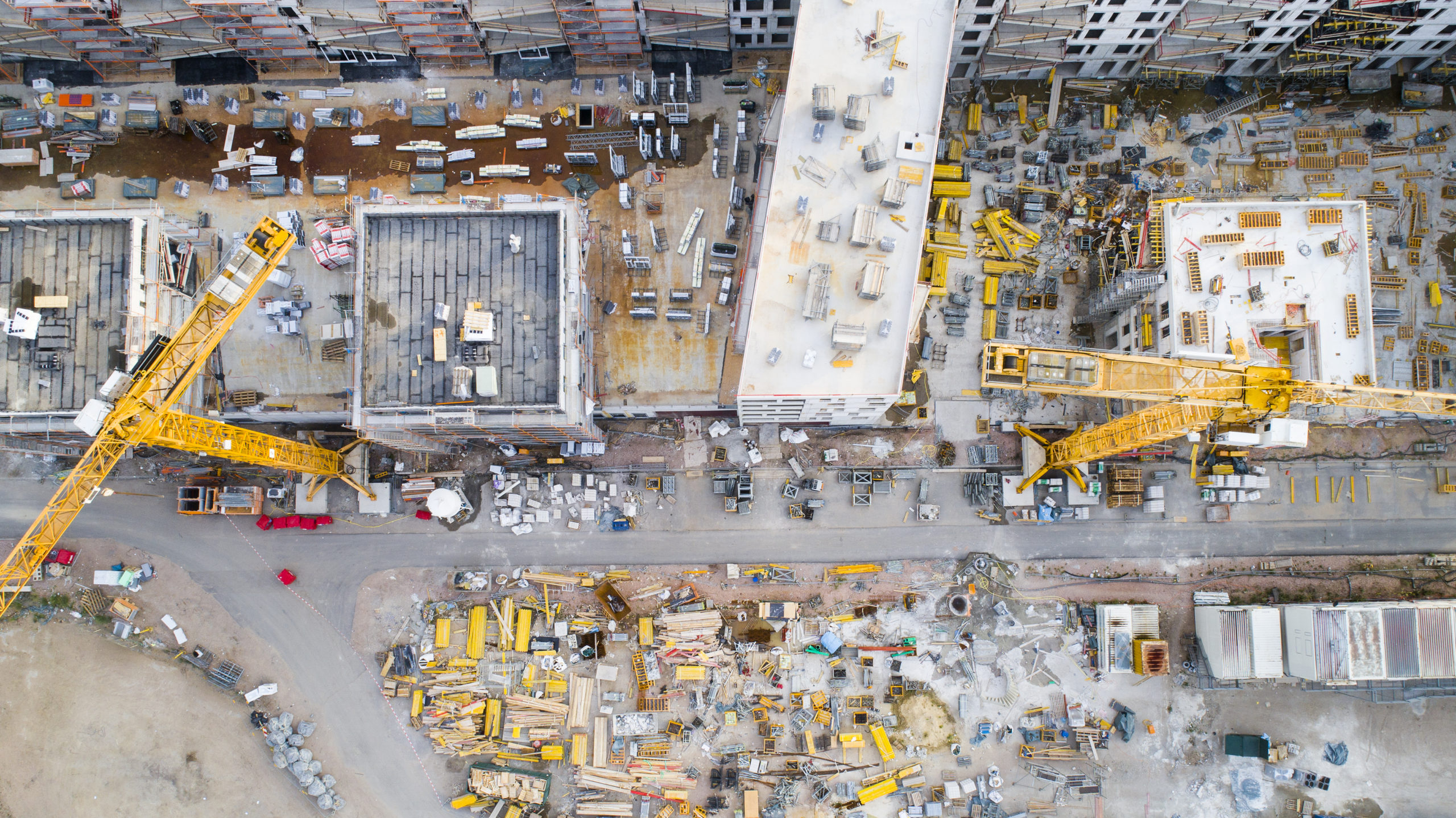 An aerial view of a construction worksite shows two yellow cranes among a sea of buildings and materials.