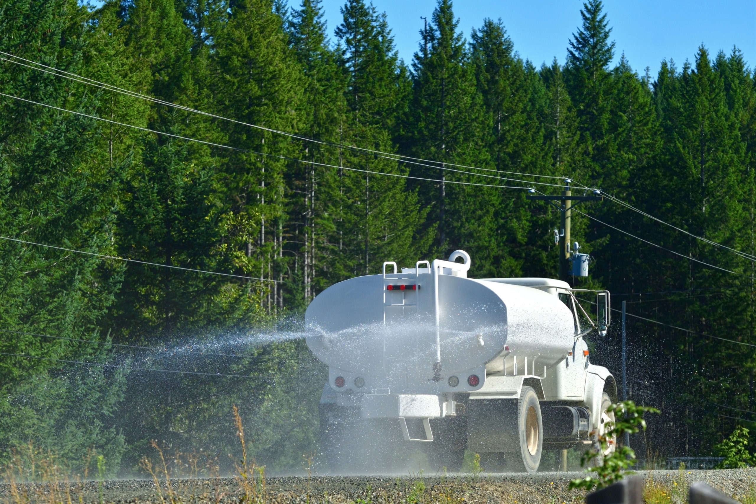 A dust suppression truck is traveling through a road surrounded by green trees while spraying water to suppress dust.