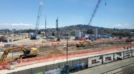 Two blue cranes and two other construction vehicles continue work at the worksite for The Walsh Group Ltd. against a bright blue sky while workers focus on concrete compressive strength monitoring..