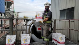 A construction worker is standing on a platform behind a ready-mix truck and holding a bag of the integral concrete hardening admixture, Hard-Cem, while being surrounded by several more bags that are lying on the ground.