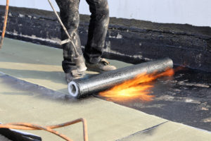 A construction worker is waterproofing a flat roof with a bitumen-sealing membrane.