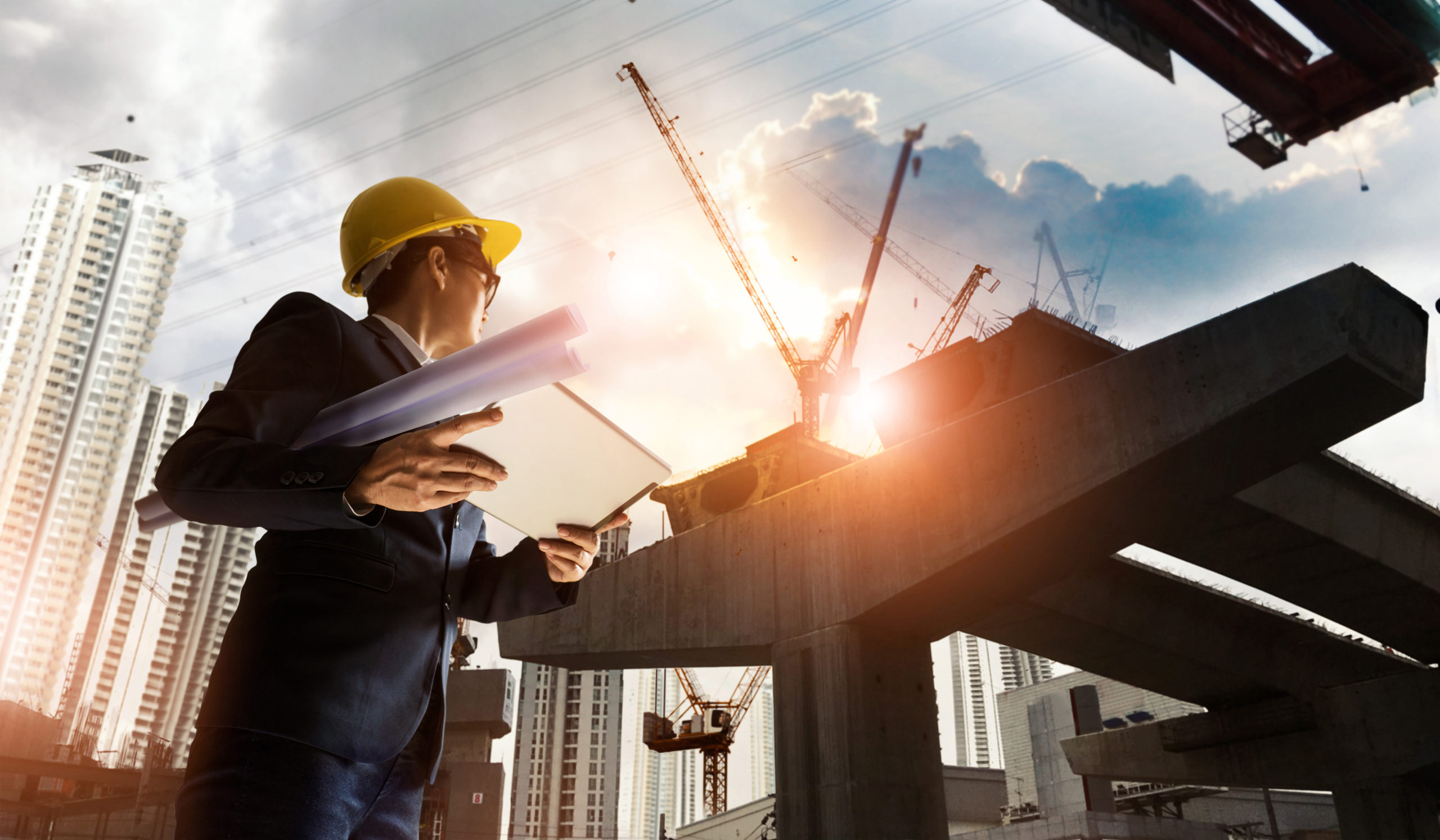 A man in a business suit and construction hat is looking up to a concrete structure near multiple cranes that is being highlighted by the sun.