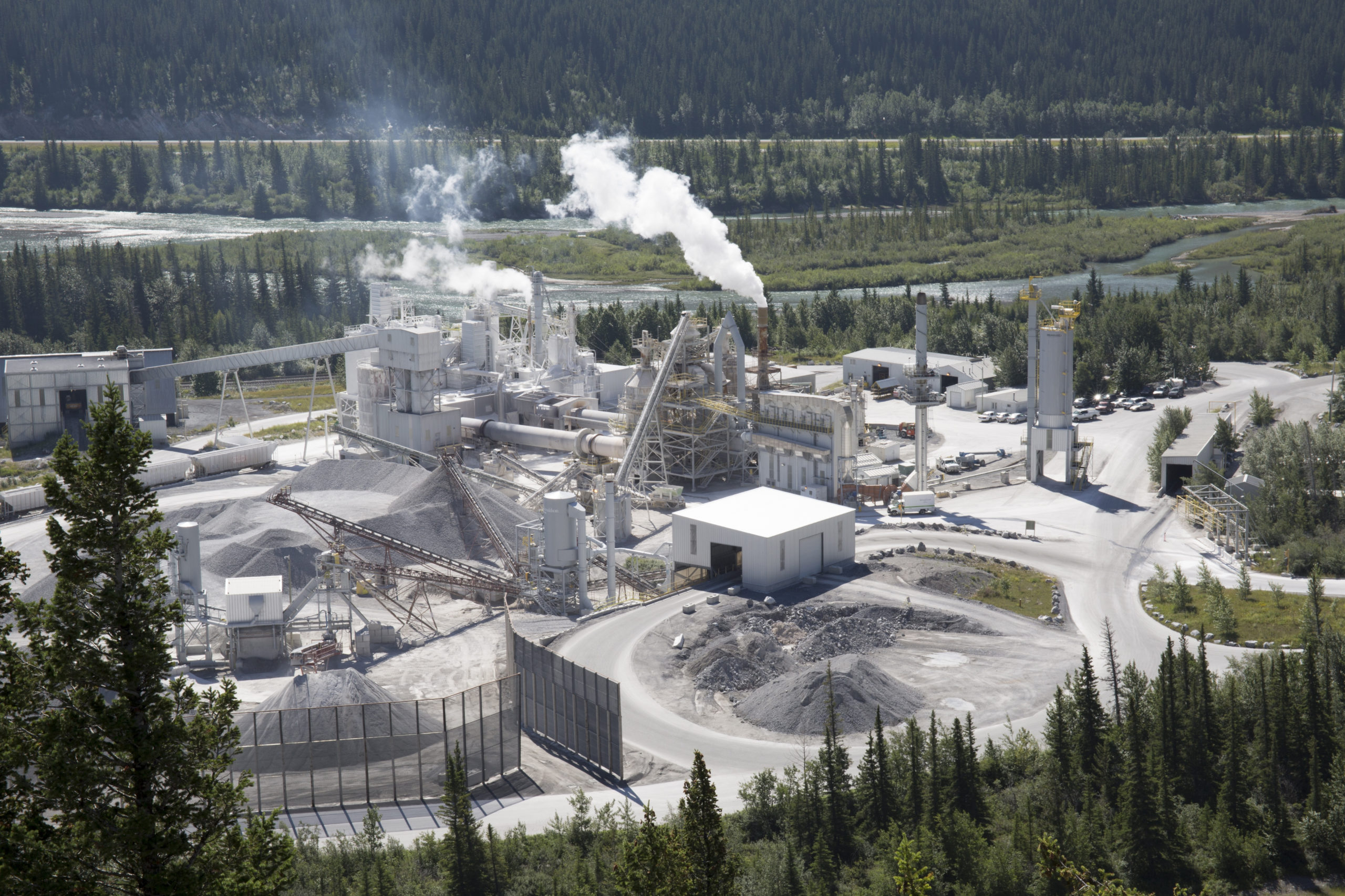 A cement manufacturing plant is sending up plumes of smoke in broad daylight surrounded by green grass and trees.