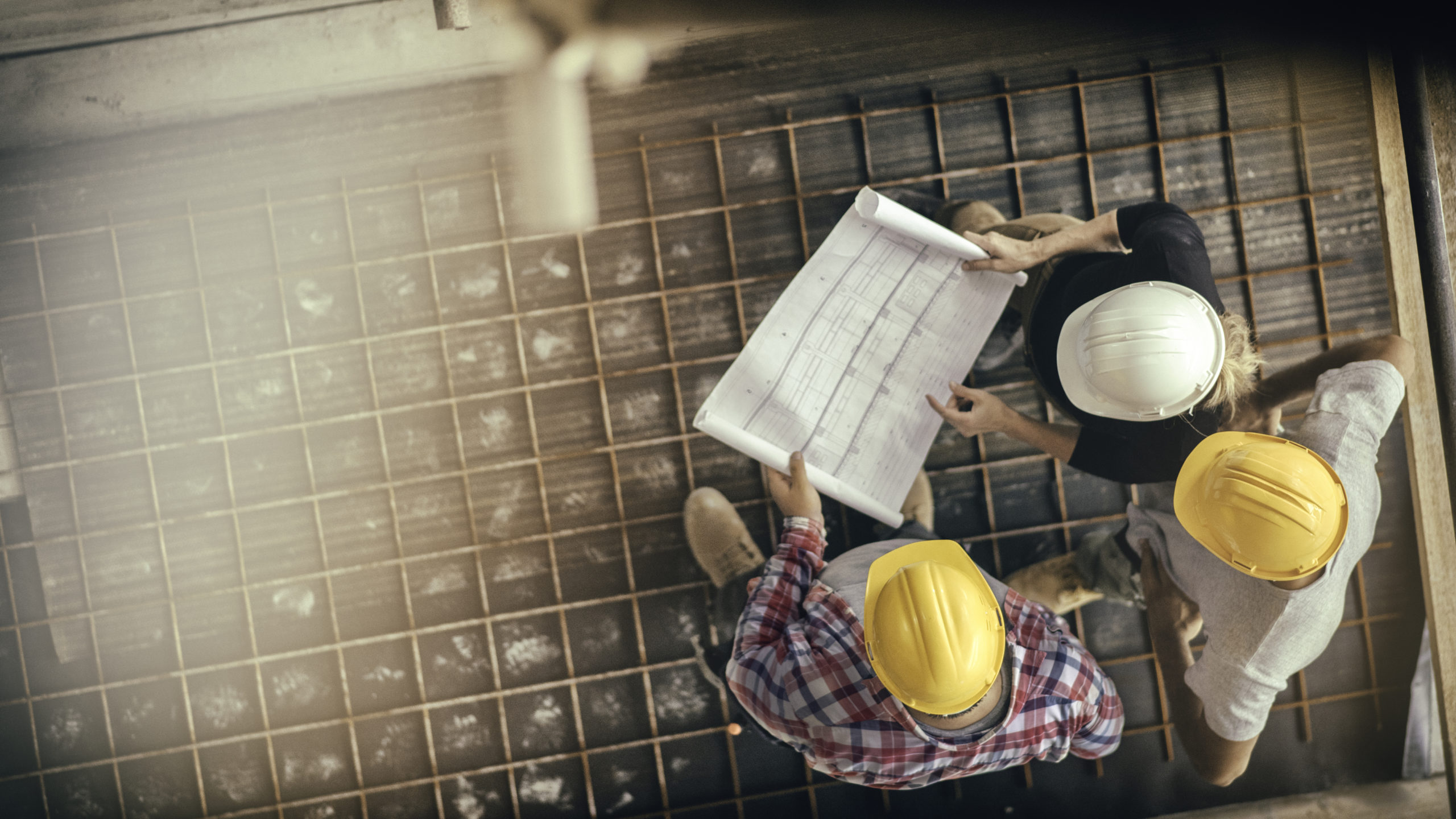 A high-angle view of three people with helmets, a female architect, foreman, and engineer, on a construction site, looking down at a blueprint.