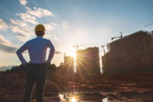 A construction worker has his hands on his hips, standing proudly as he looks on at two buildings under construction with the sun shining over one of them.