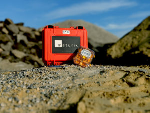 At the top of a small mound under a blue sky, a Maturix Sensor is resting at an angle against an orange carrying case with the Maturix logo on it.
