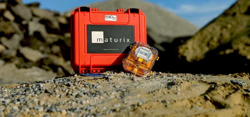At the top of a small mound under a blue sky, a Maturix Sensor is resting at an angle against an orange carrying case with the Maturix logo on it.