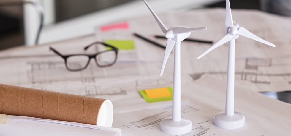 Two miniature white turbines sit on top of architecture plans for Passive House and net-zero construction next to a cardboard tube, a pair of glasses, and two pencils.