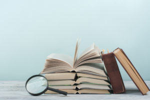 A magnifying glass rests against a pile of books laying open-faced that have two closed books leaning sideways onto them.