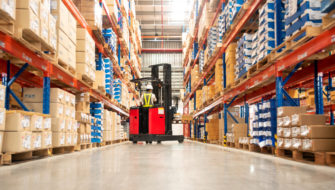 A worker in a forklift is loading packed goods onto shelves in a packed warehouse that's been designed with warehouse optimization in mind.