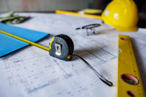 A rectractable measuring tape has been pulled out near safety glasses, a hard hat, and paper. All of which are lying across blueprints for a construction project that will use circular economy solutions.