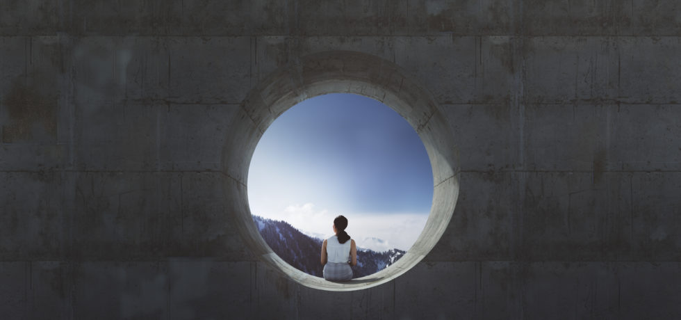 A young woman is sitting in a circular concrete opening and looking out at mountains.