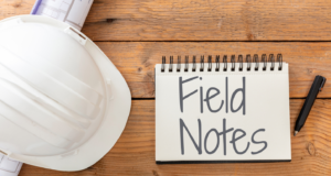 On a wooden table, a white hard hat is resting on top of blue prints next to a note pad with the title "Field Notes" on it, which is to the left of a black pen.