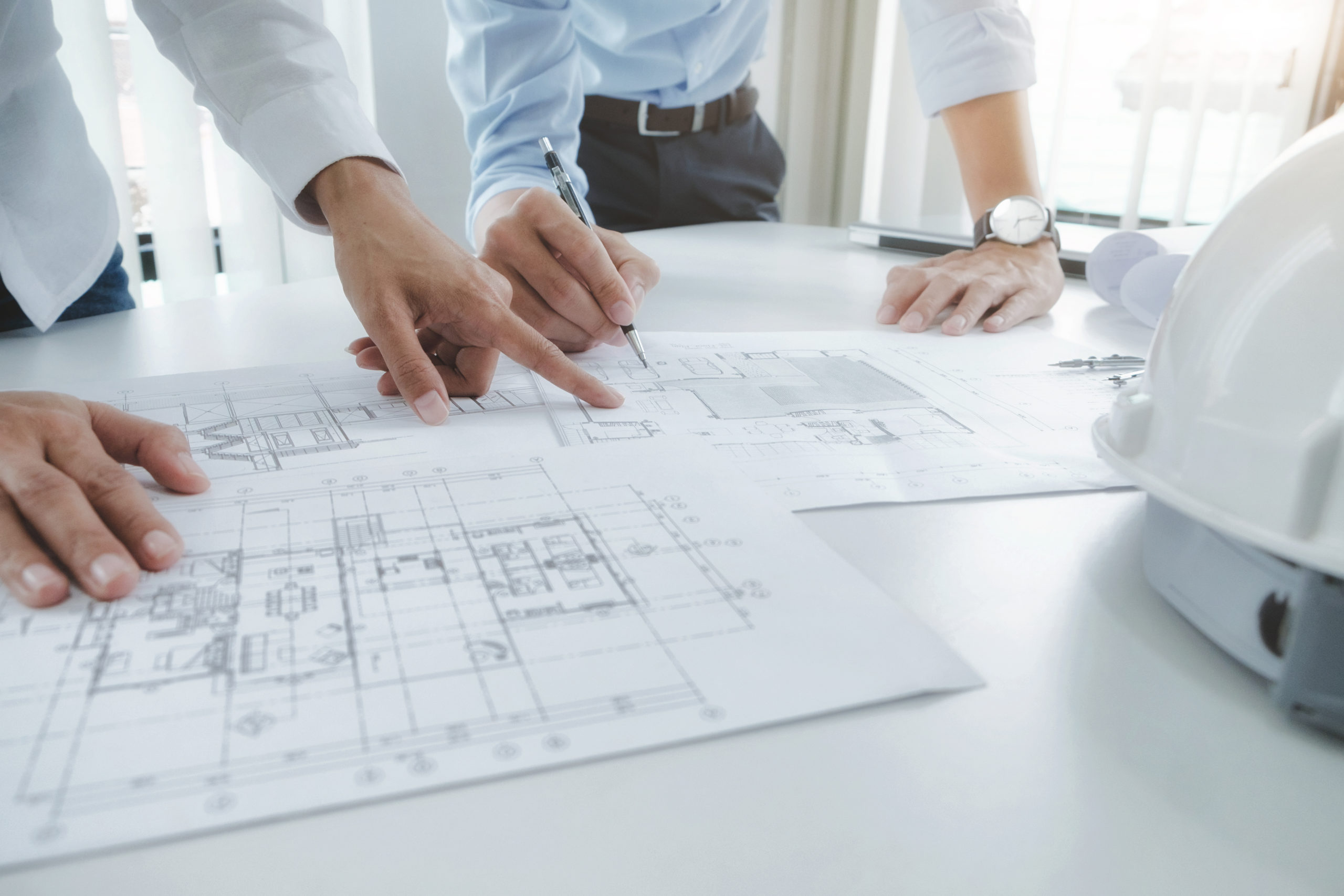 One hand is pointing at the designs for a building on white paper while another set of hands writes on a different sheet of plans.