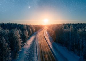 A concrete road surrounded by snowy trees is leading towards a sunset with stars from the Milky Way showing above it.