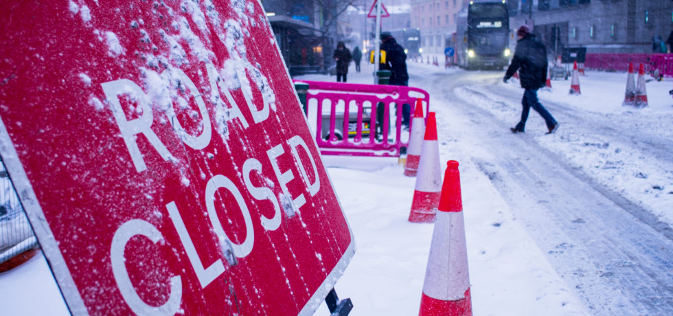 A sign viewed from its side says "Road Closed" in snowy weather next to traffic cones, a frozen road, pedestrians walking by, and a bus arriving.