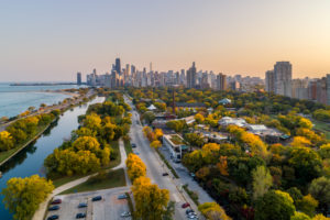 An aerial view of Lincoln Park in Chicago shows bodies of water to the left and groups of beatiful green and yellow trees separated by strips of pavement, which leads to the Chicago skyline in the background.