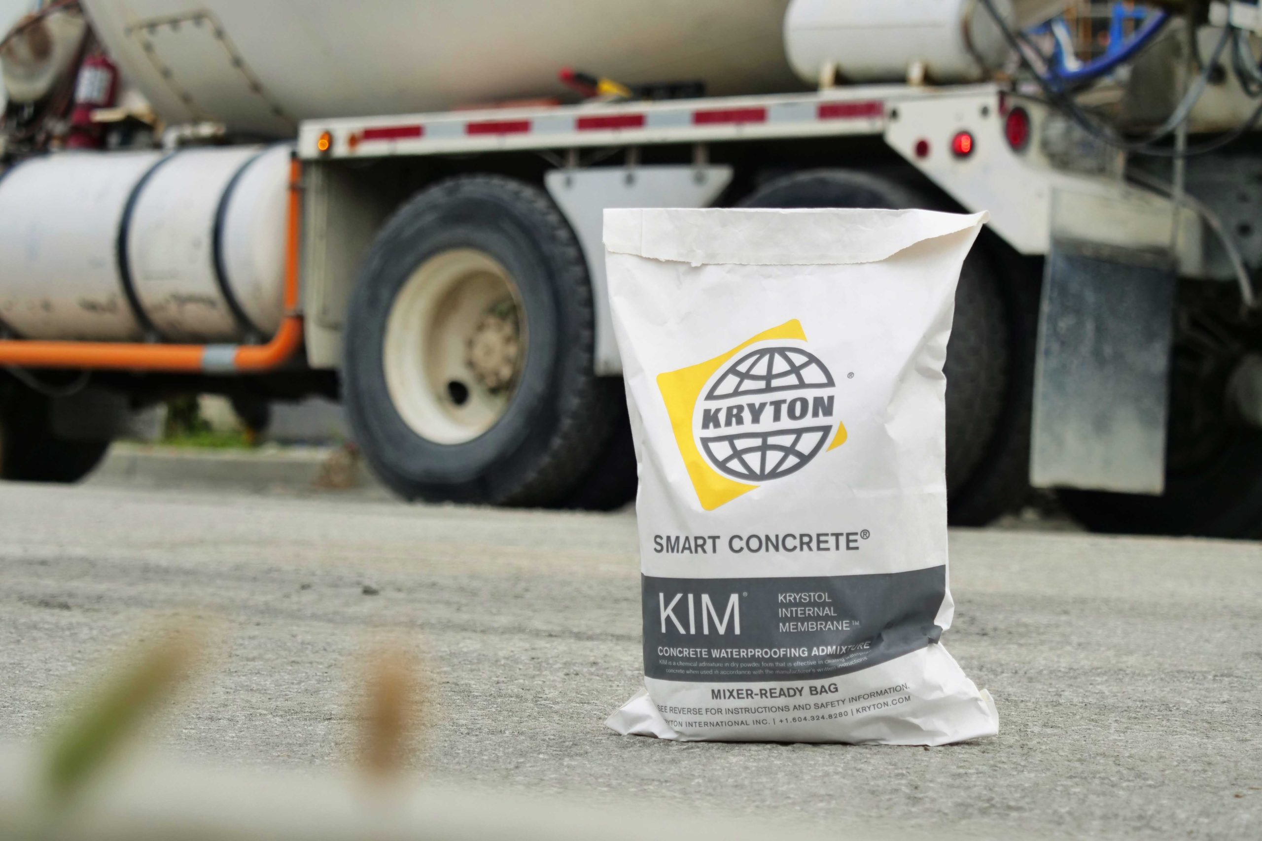 A dissoluble bag of KIM is sitting on pavement ahead of a ready-mix truck that's parked off to the side.